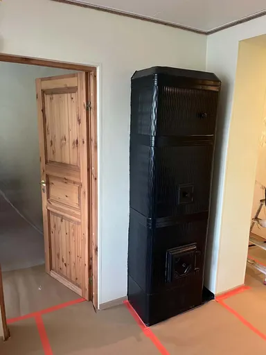 Black sheet-metal stove with cast-iron doors in the corner of a room next to a wooden door, with the floor covered and taped