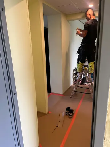 Corridor with a paper-covered floor and a worker on a ladder putting up plastic sheeting and tape