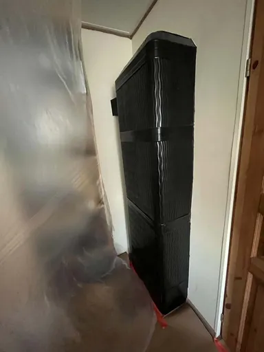 Stove with a black sheet-metal casing in the corner of a room, with an area covered in plastic sheeting beside it and the floor covered in paper